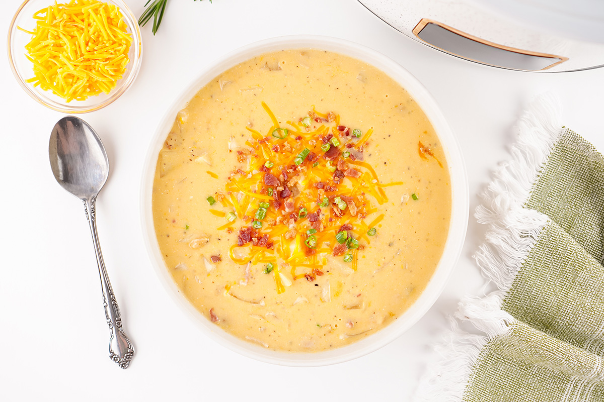 Overhead view of a bowl of crock pot crack potato soup topped with shredded cheddar, crumbled bacon, and chopped green onions next to a spoon and green napkin.