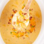 Wooden spoon lifting a hearty scoop of crock pot crack potato soup filled with potatoes, cheddar cheese, bacon, and green onions from a slow cooker.
