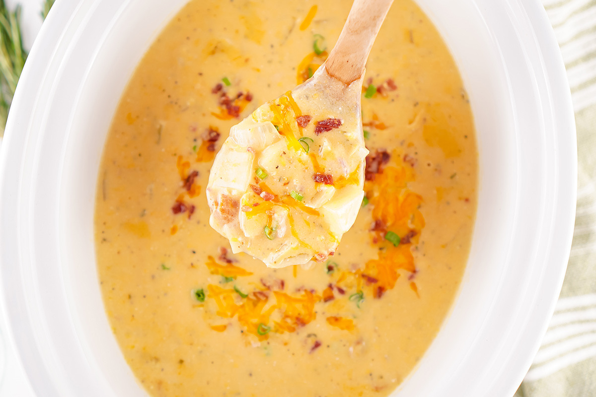 Wooden spoon lifting a hearty scoop of crock pot crack potato soup filled with potatoes, cheddar cheese, bacon, and green onions from a slow cooker.