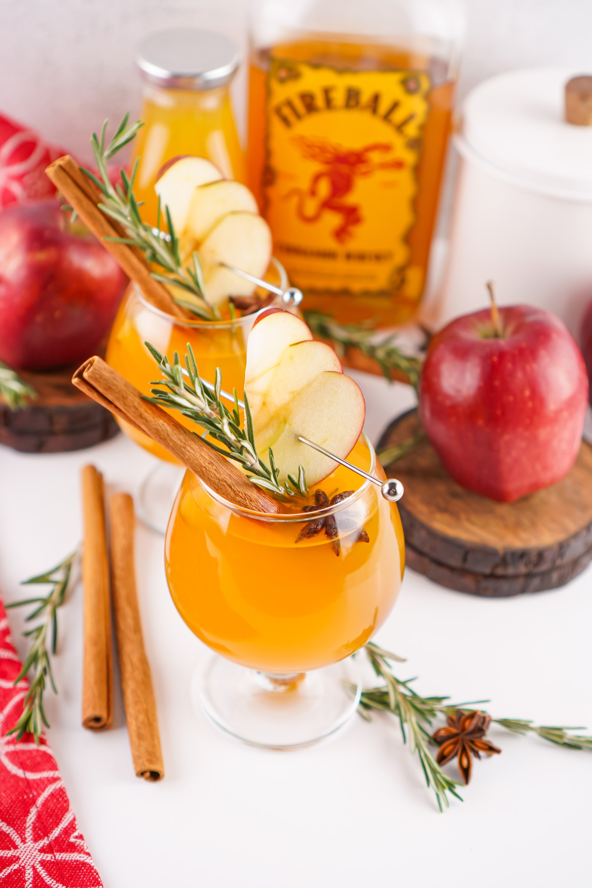 Two cocktails made with apple cider and Fireball whiskey served with apple slices, rosemary, and cinnamon sticks surrounded by fresh apples.