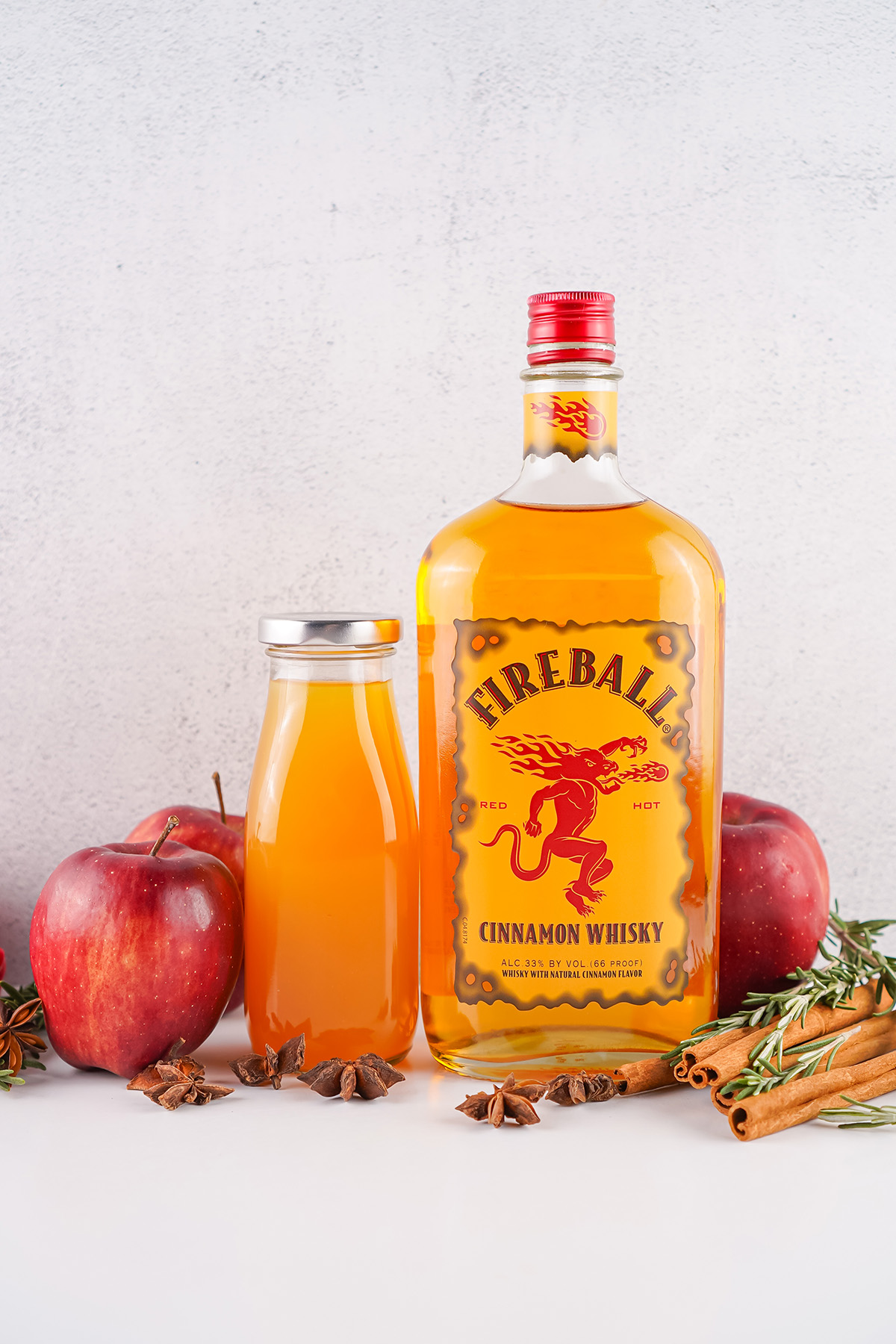 Bottle of Fireball whiskey next to fresh apples, cinnamon sticks, and a small jar of apple cider. 