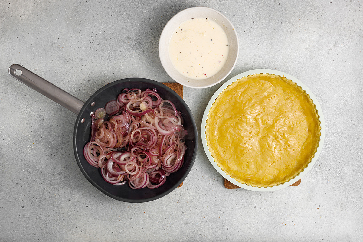 Flaky pastry, sweet caramelized onions and a creamy egg and Gruyere cheese filling make this onion tart both flavorful and irresistible to serve as an appetizer, dinner or even brunch. Caramelized onions in a skillet beside a bowl of cream and eggs, and the pre-baked tart crust.