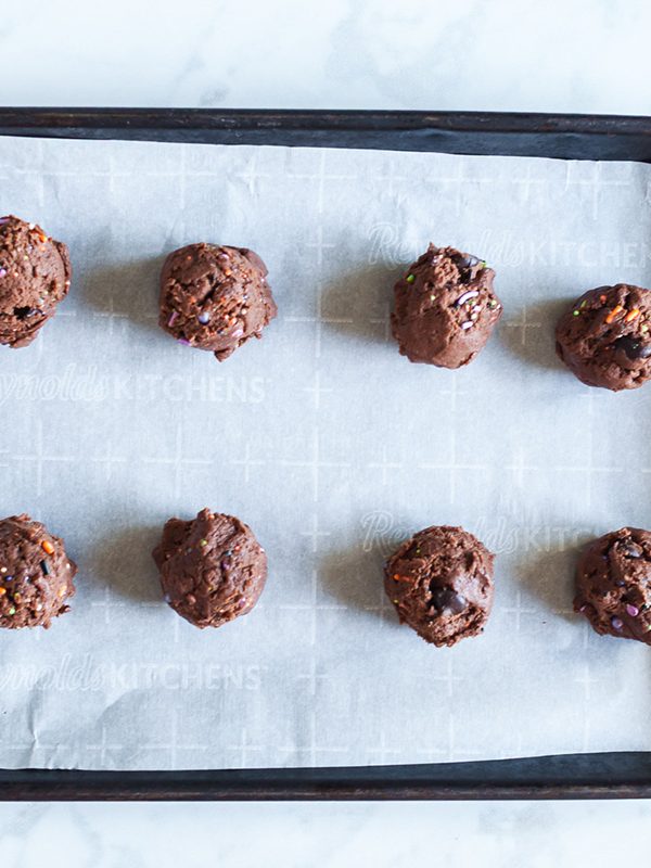 Scoops of chocolate cookie dough with sprinkles on a parchment-lined baking sheet before baking