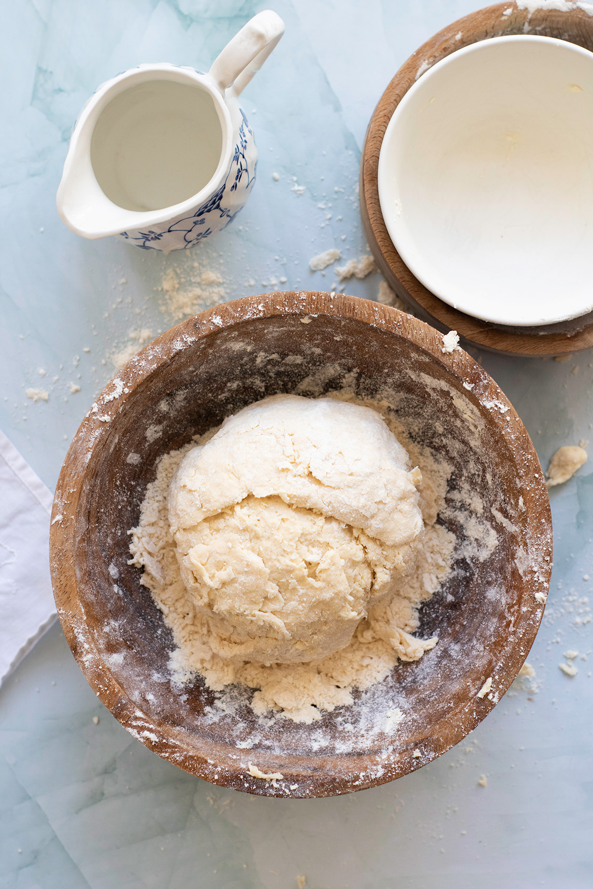 Pie dough ball in a floured wooden bowl with a small pitcher of water nearby