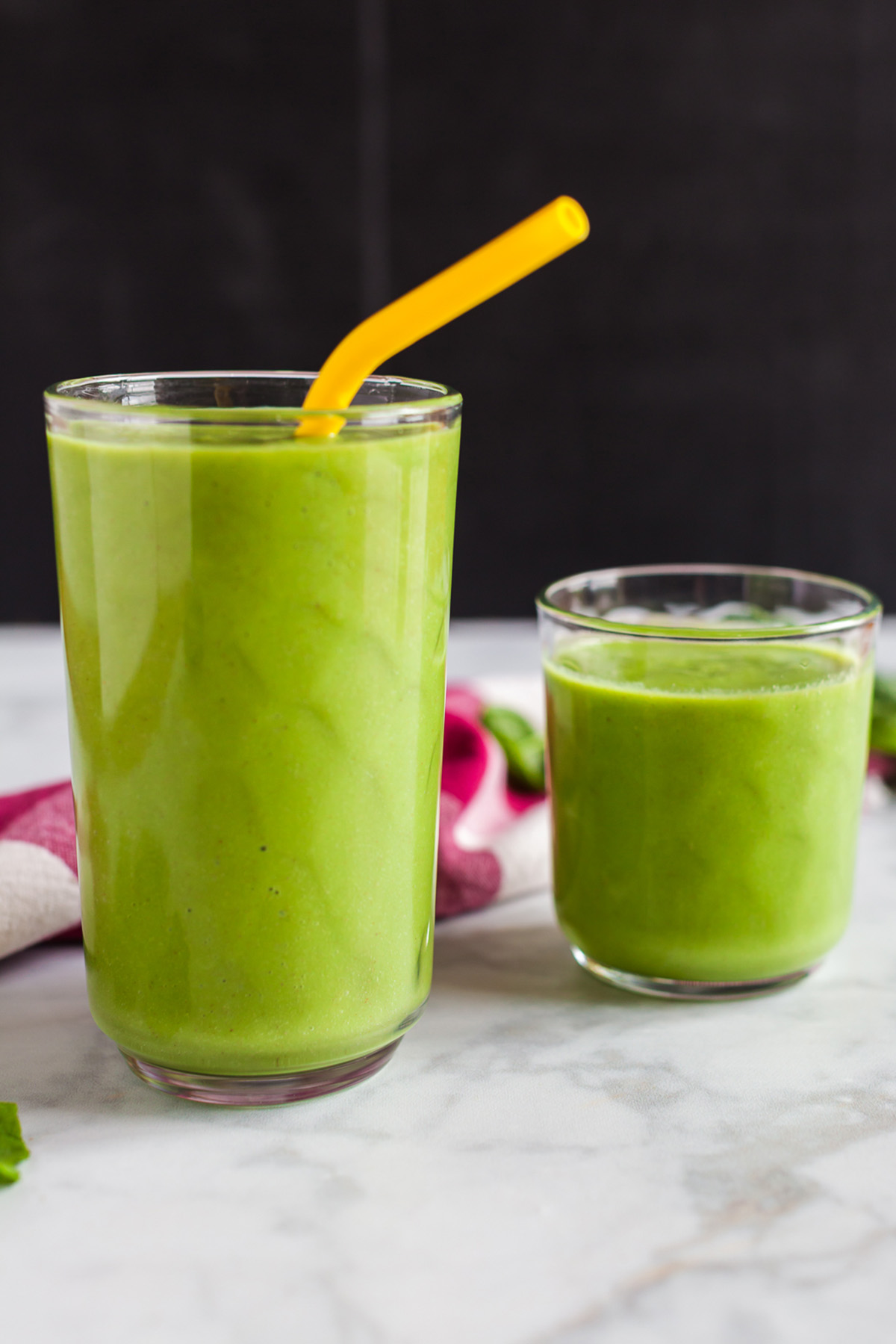 Side view of a tall and short glass filled with spinach green apple smoothie, with a yellow straw in the taller glass.