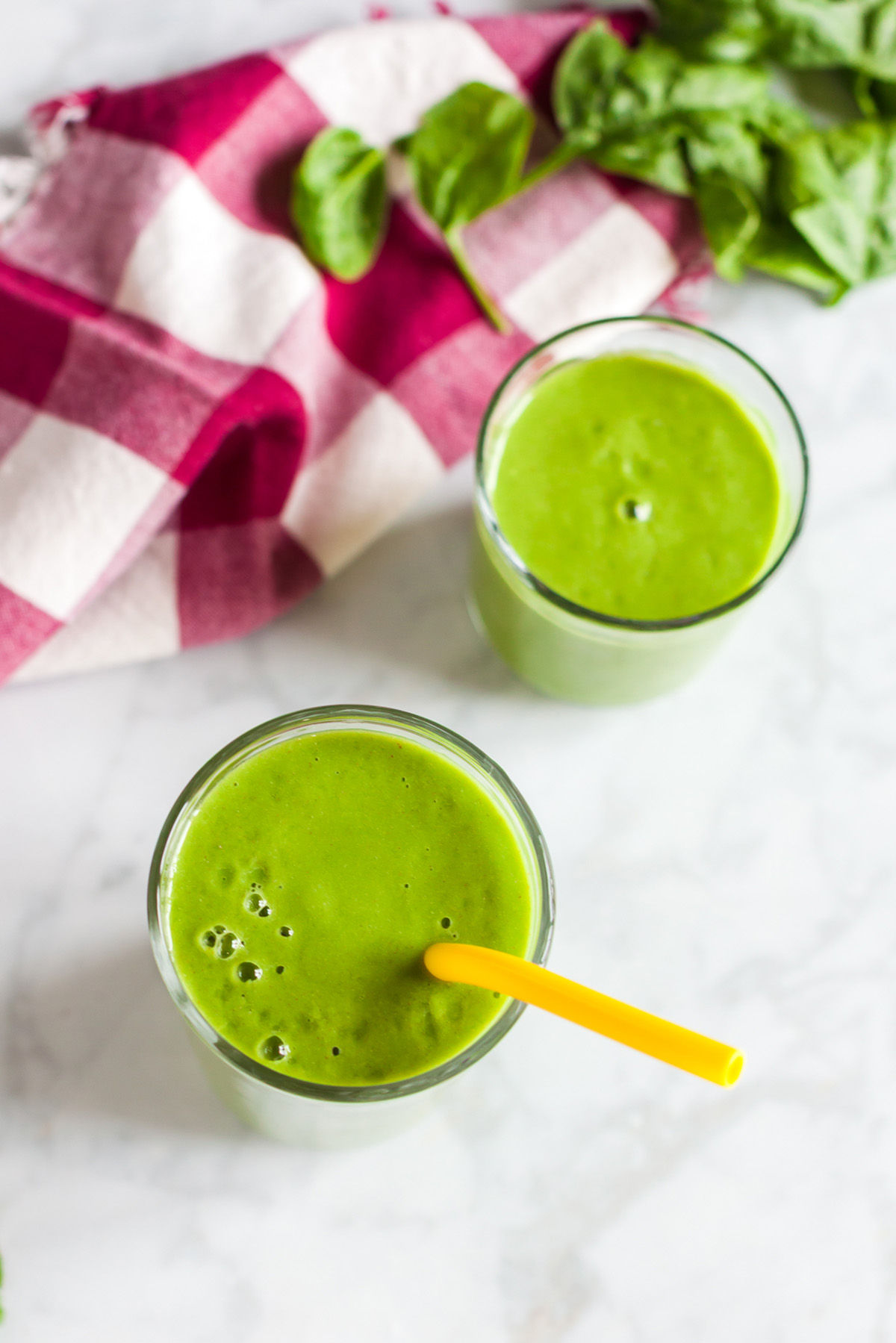 Overhead photo of two spinach apple smoothies on a marble surface with spinach leaves around.