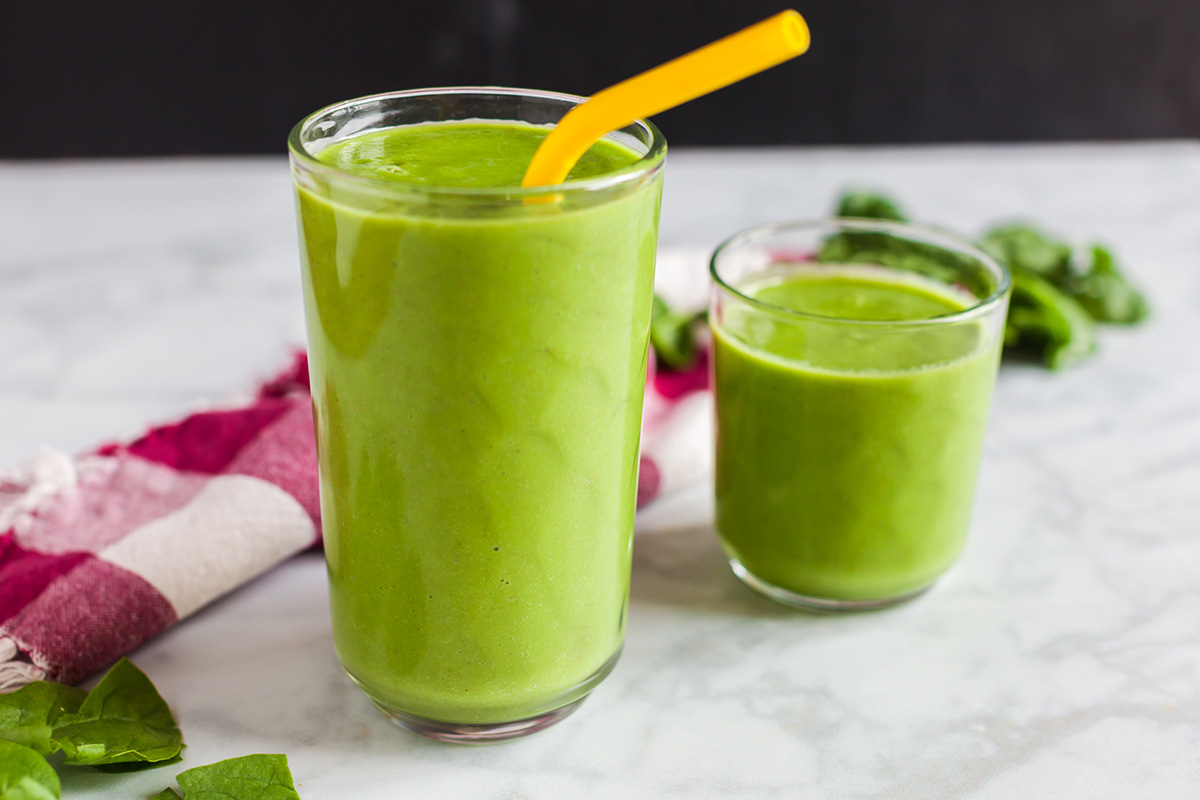 Horizontal view of spinach apple smoothies in two glasses, garnished with a yellow straw in one.