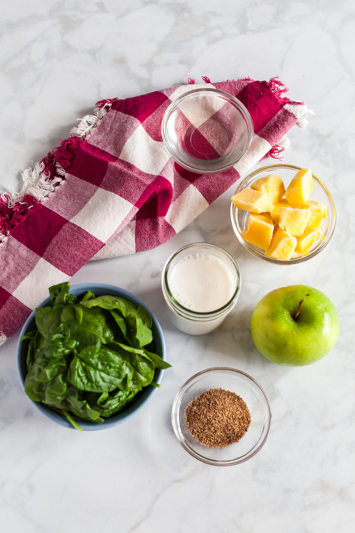 Overhead photo of spinach green apple smoothie ingredients including fresh spinach, a green apple, frozen mango chunks, ground flaxseed, and almond milk on a marble surface with a red checkered napkin.
