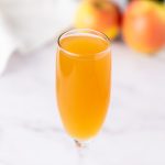 Close-up of a bubbly apple cider mimosa in a glass, with fresh apples and a bottle of champagne blurred in the background.