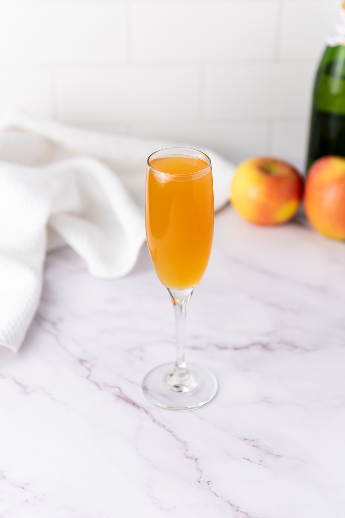 A champagne flute filled with golden apple cider mimosa on a marble countertop with fresh apples and a white towel in the background.