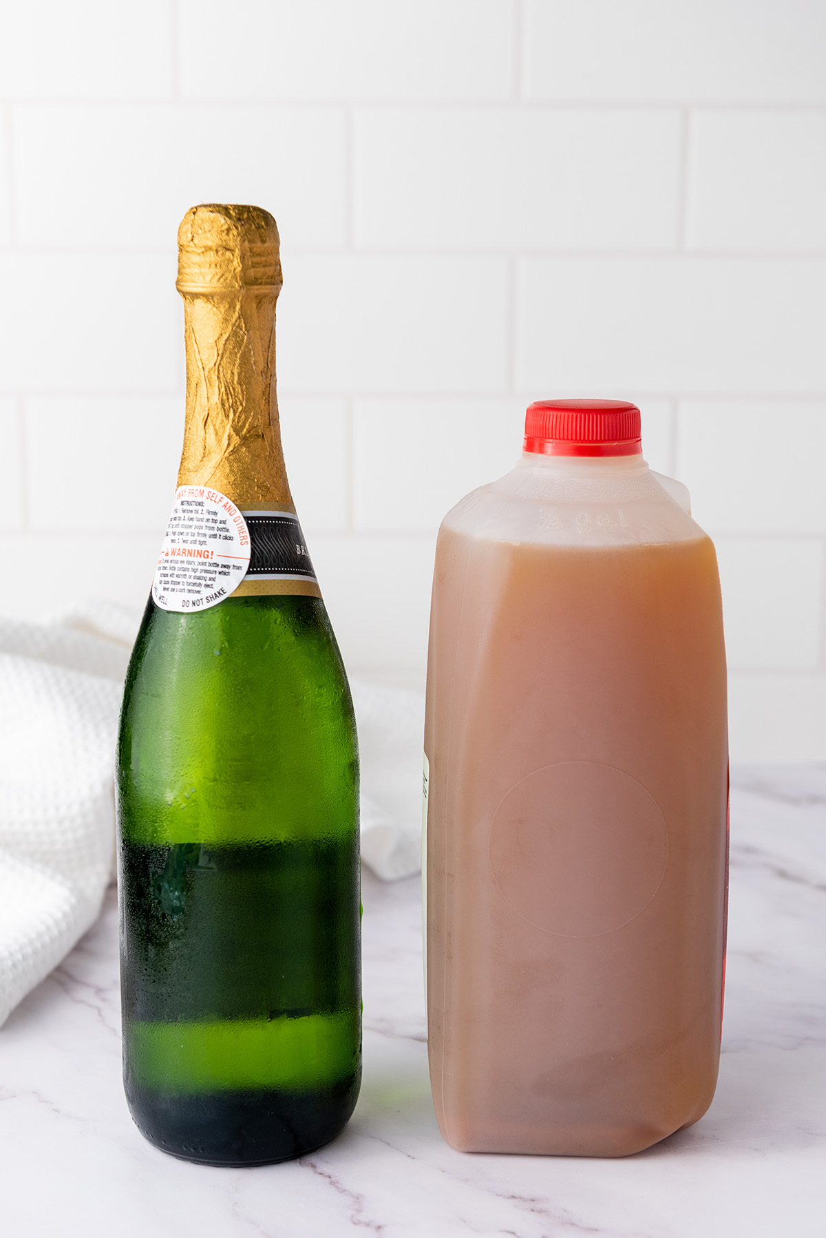 A glass of apple cider mimosa on a marble surface with a jug of apple cider and a champagne bottle in the background.