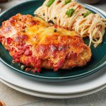 Close-up of golden chicken Parmesan baked with marinara and melted cheese, plated with spaghetti tossed in tomato sauce and fresh basil leaves.