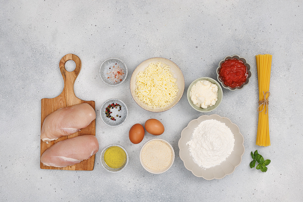 Flat lay of chicken Parmesan ingredients including chicken breasts, eggs, flour, breadcrumbs, Parmesan, mozzarella, marinara sauce, and spaghetti.