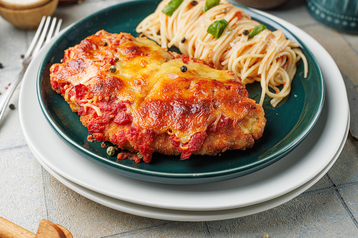 Close-up of golden chicken Parmesan baked with marinara and melted cheese, plated with spaghetti tossed in tomato sauce and fresh basil leaves.