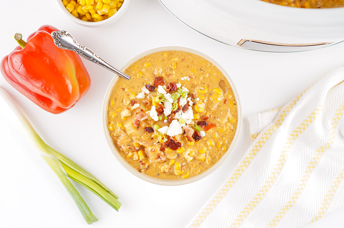 Overhead view of a bowl of creamy slow cooker Mexican street corn soup topped with bacon, queso fresco, and green onions, surrounded by fresh bell pepper, green onions, and a yellow-striped napkin.