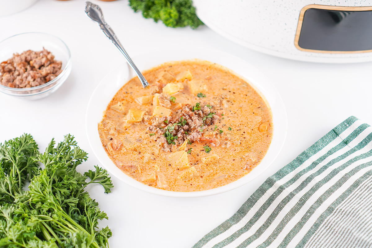 Bowl of slow cooker sausage potato soup topped with crumbled sausage and parsley, served next to fresh herbs.