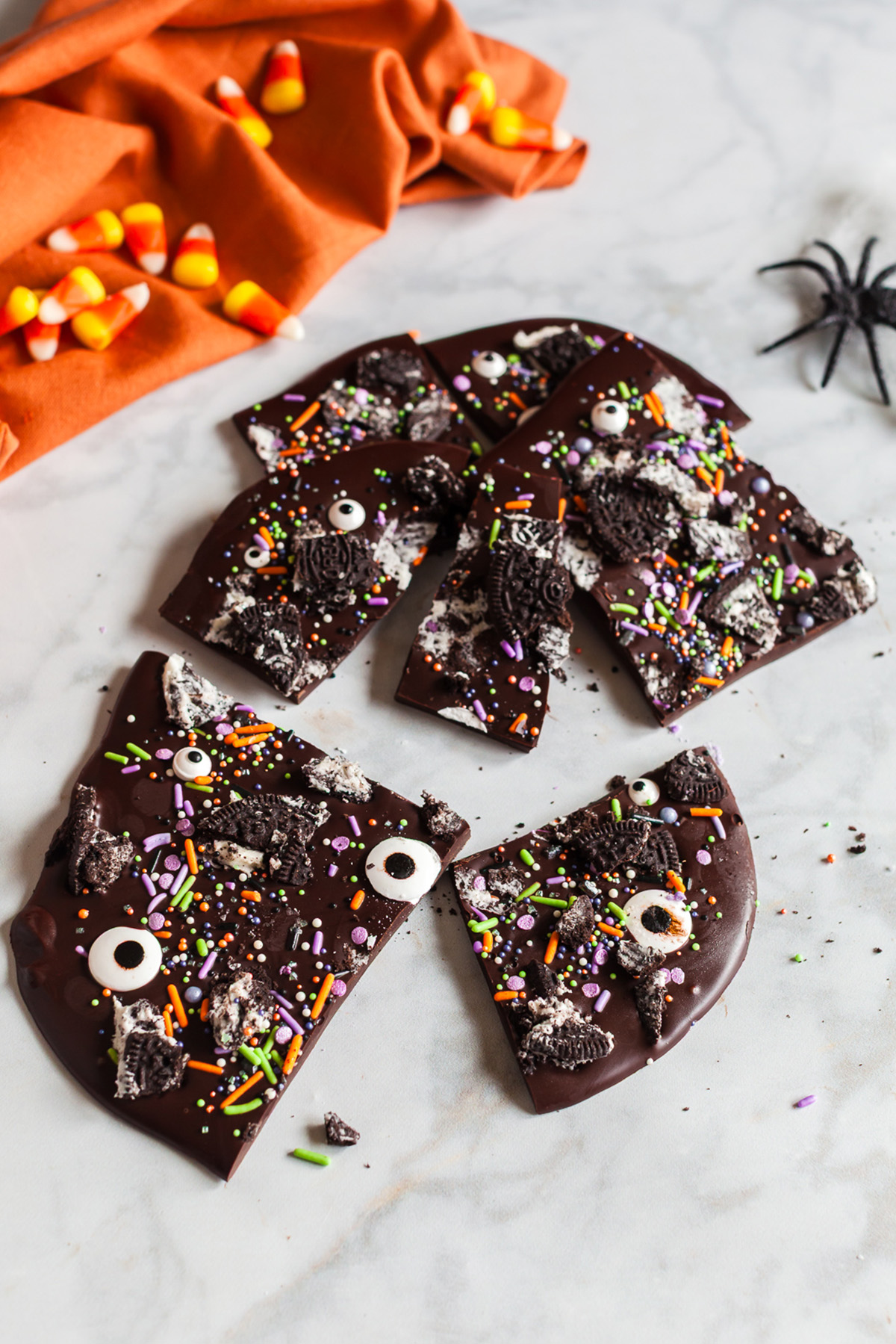 Dark chocolate Halloween bark pieces on a marble surface with orange cloth, candy corn, and toy spiders for decoration.