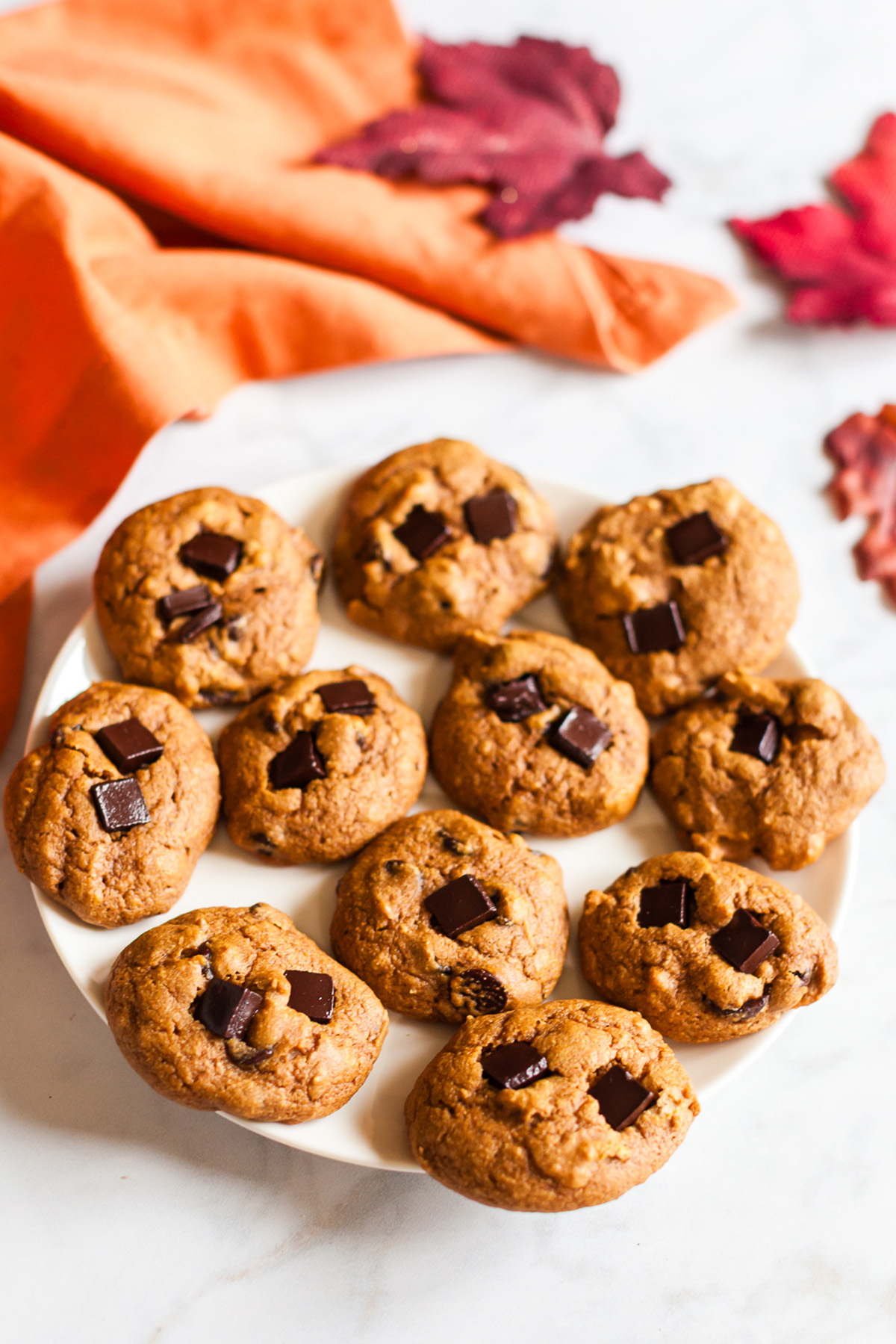 Plate filled with chewy pumpkin spice breakfast cookies topped with chocolate chunks, surrounded by orange napkin and fall leaves.