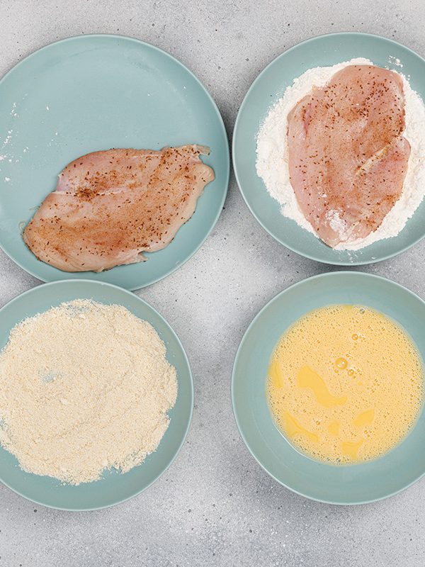 tep-by-step of breading chicken for chicken Parmesan—seasoned chicken breasts, flour, beaten eggs, and breadcrumbs on separate plates.