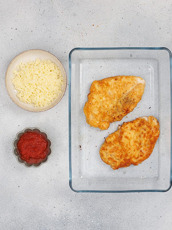 Baked chicken breasts in a dish with bowls of marinara sauce and shredded mozzarella ready to assemble chicken Parmesan.