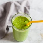 Smooth and creamy green pear smoothie with yellow straw on marble counter, gray napkin in background.