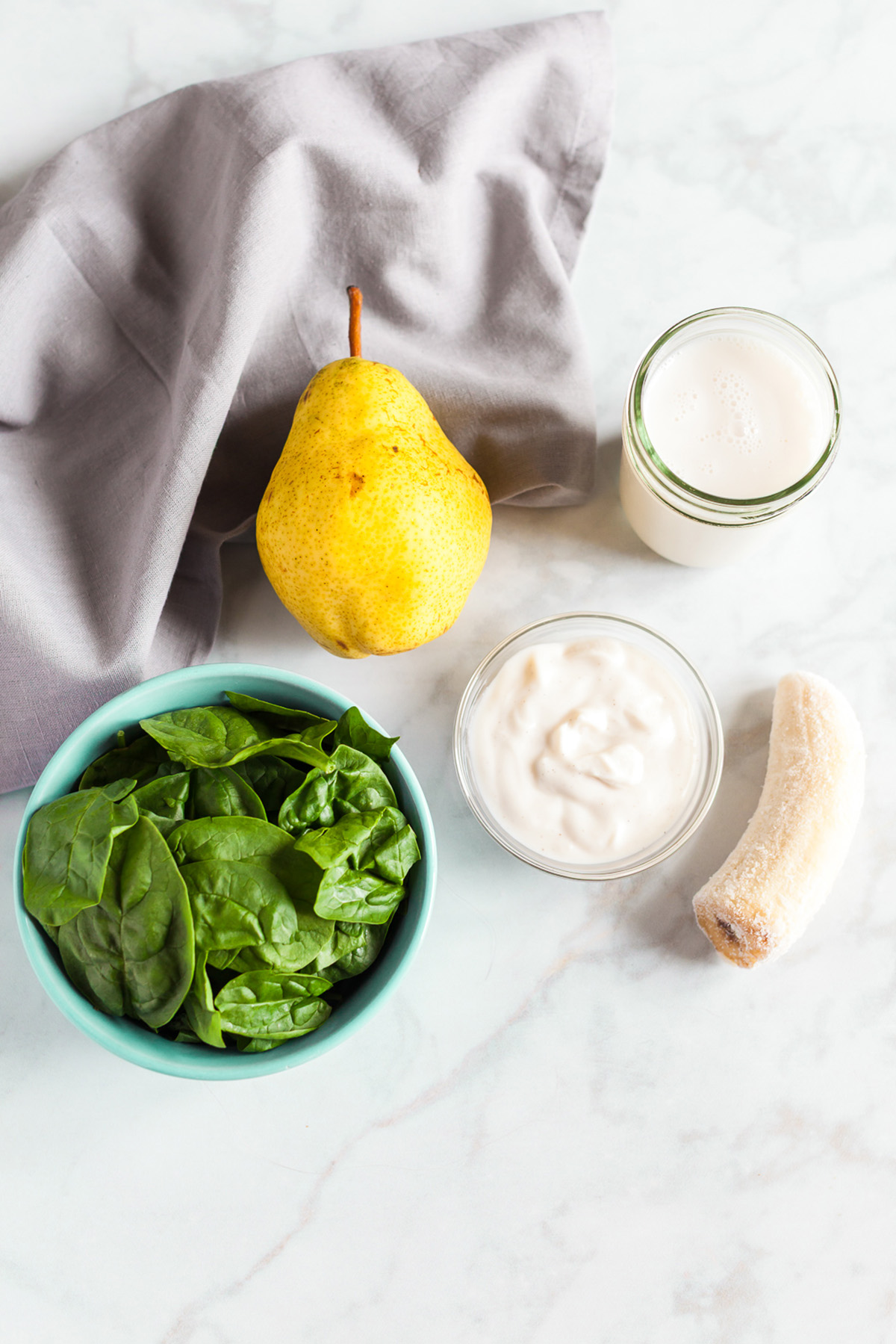 Ingredients for pear smoothie, fresh spinach, ripe pear, banana, yogurt, and milk arranged on marble countertop.