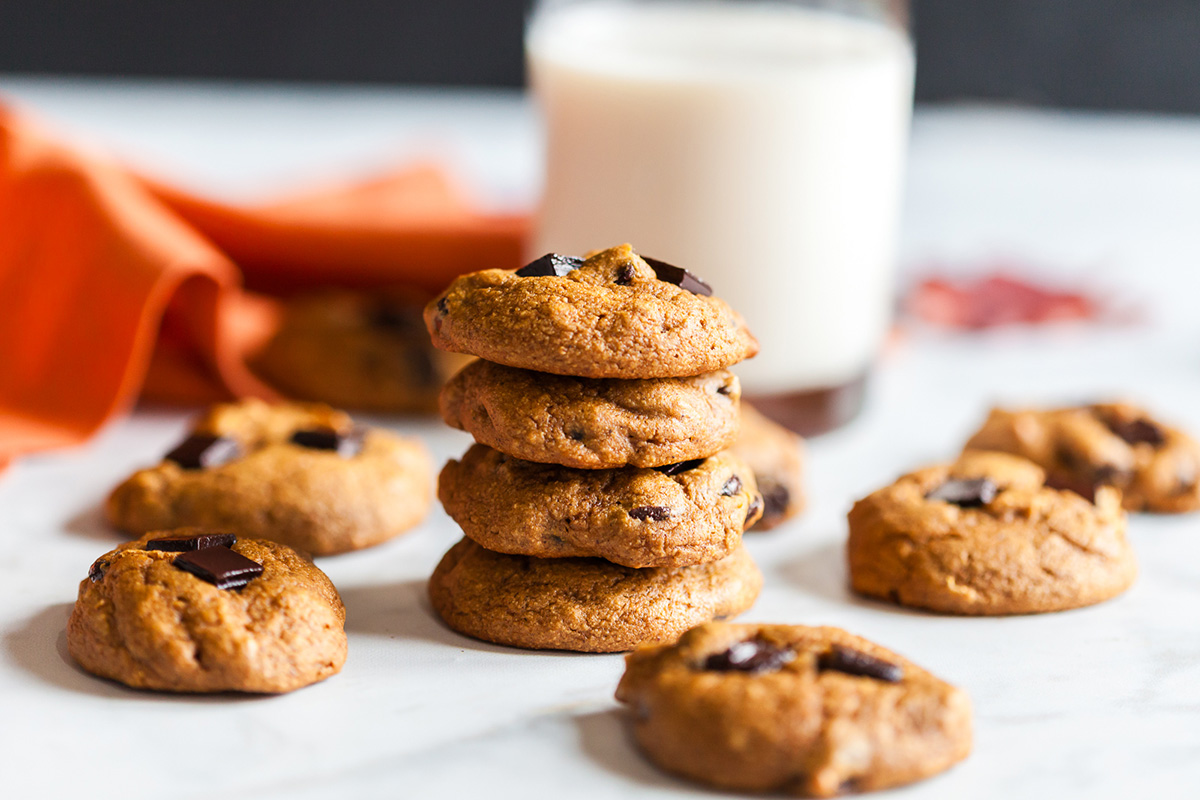 Stack of soft pumpkin oat breakfast cookies with dark chocolate chunks beside a glass of milk and an orange napkin, a cozy fall breakfast treat.