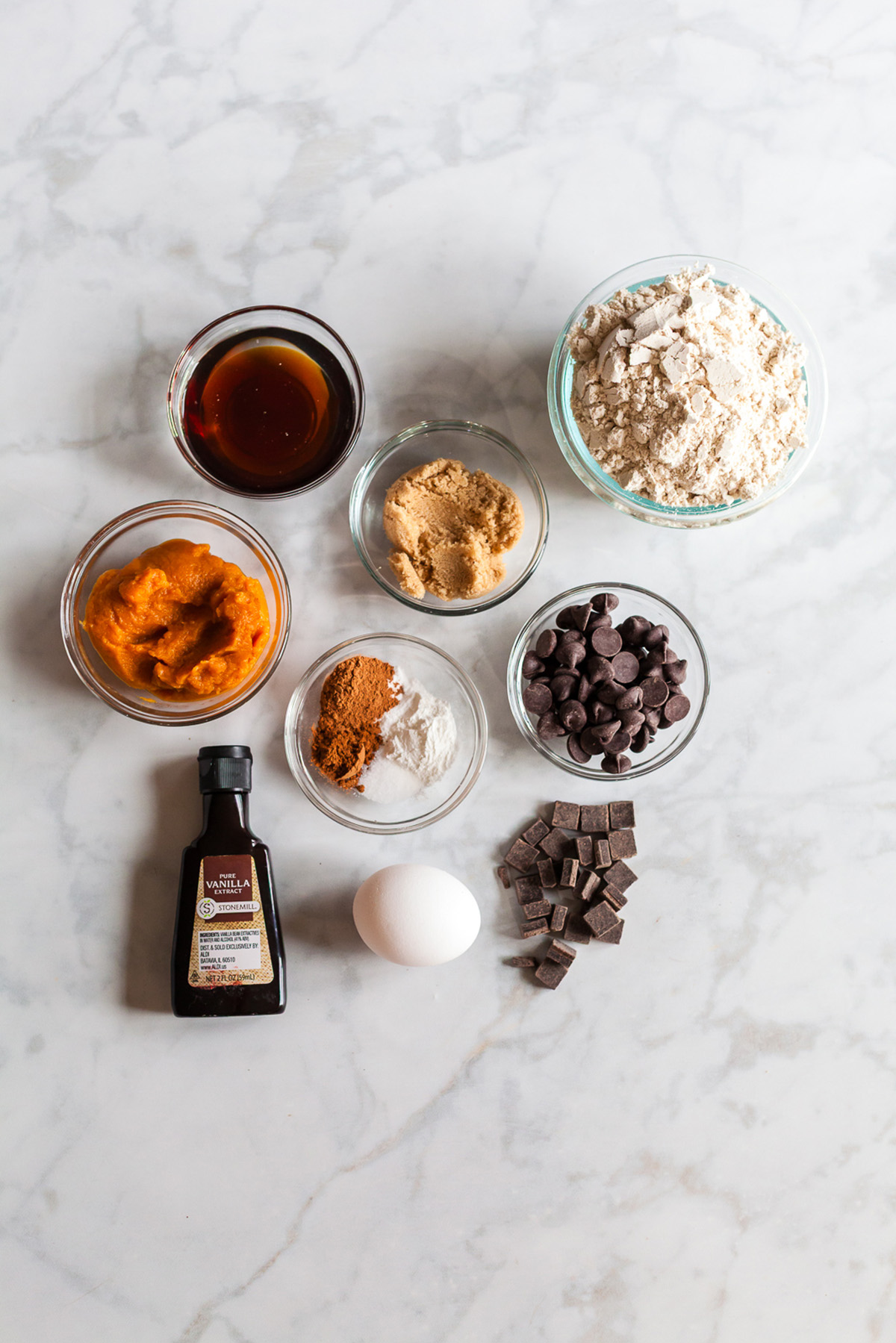 Glass bowls filled with oat flour, pumpkin puree, brown sugar, maple syrup, spices, chocolate chips, vanilla, and an egg on a marble surface, ingredients for pumpkin oat breakfast cookies.