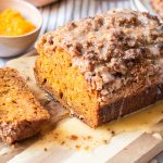 Loaf of pumpkin streusel bread topped with glaze and sliced to reveal its tender crumb, with fresh squash and pumpkin puree in the background.