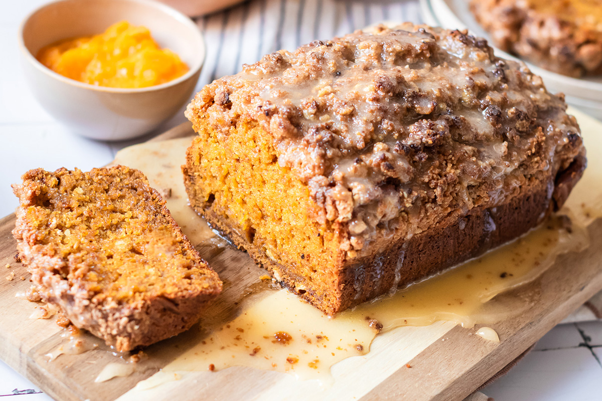 Loaf of pumpkin streusel bread topped with glaze and sliced to reveal its tender crumb, with fresh squash and pumpkin puree in the background.