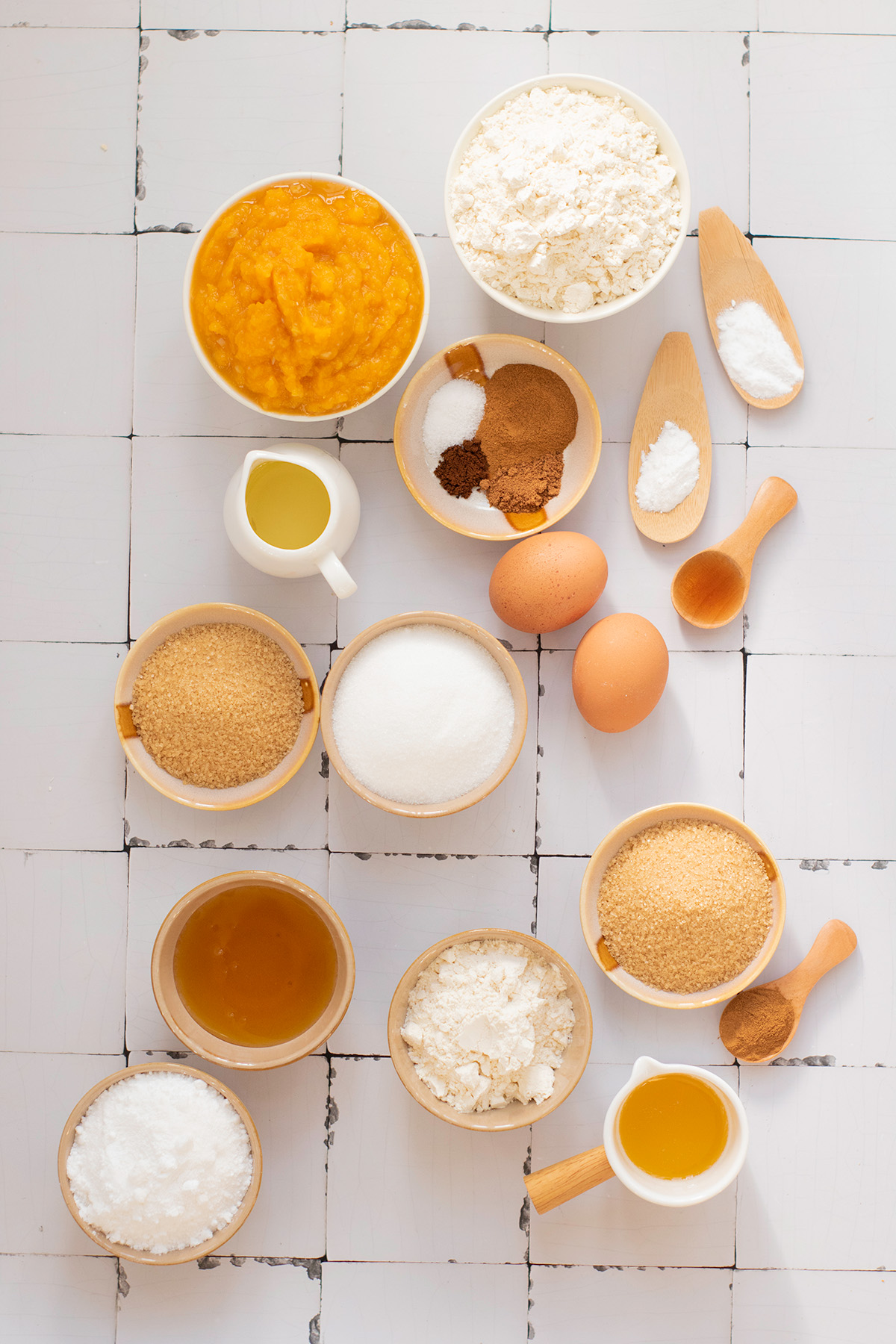 Overhead view of ingredients for pumpkin streusel bread, including flour, eggs, pumpkin puree, brown sugar, oil, honey, and warm spices arranged neatly on a white tiled surface.