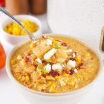 Bowl of Mexican street corn soup topped with crispy bacon, queso fresco, and green onions, with red bell pepper and corn kernels in the background.