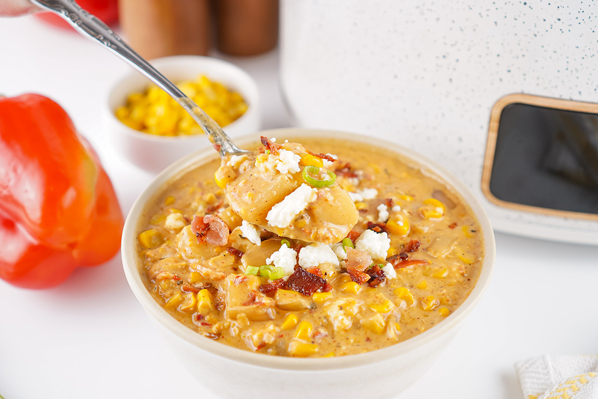 Bowl of Mexican street corn soup topped with crispy bacon, queso fresco, and green onions, with red bell pepper and corn kernels in the background.
