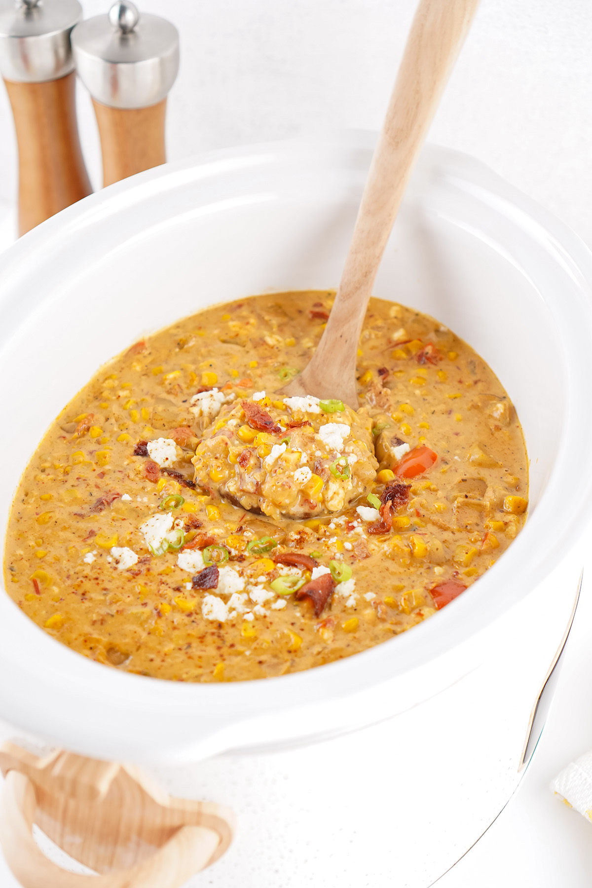 Creamy corn and potato soup simmering in a white slow cooker, topped with bacon and crumbled queso fresco, ready to serve.