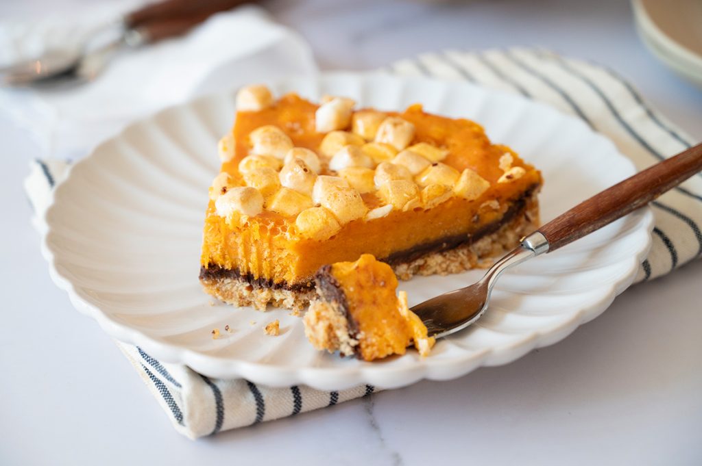 Slice of sweet potato pie with marshmallow topping on a white scalloped plate, with a fork taking a bite to show the chocolate layer and graham cracker crust.
