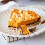 Slice of sweet potato pie with marshmallow topping on a white scalloped plate, with a fork taking a bite to show the chocolate layer and graham cracker crust.