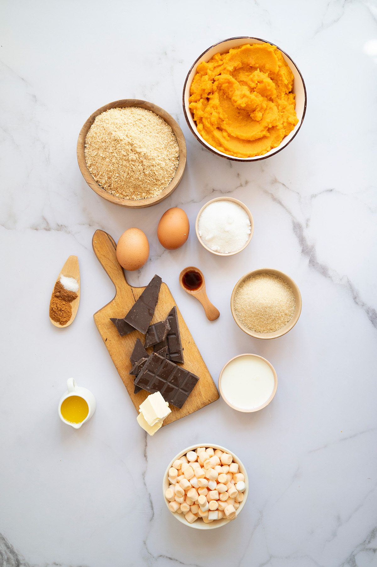 Overhead view of ingredients for sweet potato s’mores pie including mashed sweet potatoes, graham cracker crumbs, chocolate, marshmallows, eggs, cream, and spices arranged on a marble counter.