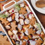 Gingerbread French toast casserole decorated with gingerbread cookies, rosemary sprigs, pecans, and pomegranate seeds, shown with a blue checkered cloth and a small pitcher of syrup.