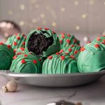 A close-up view of Grinch Oreo Balls on a silver plate, decorated with green candy drizzle and red heart sprinkles, with one truffle revealing the dark Oreo center.