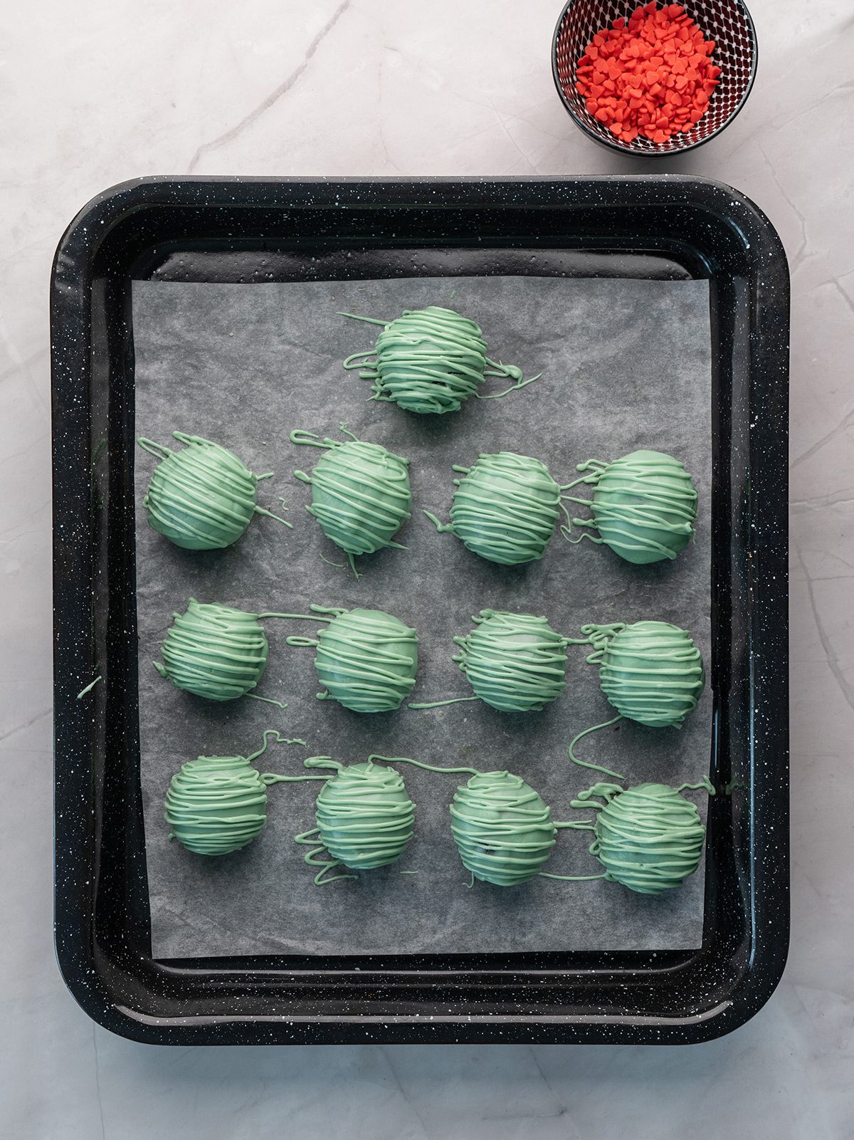 Turn classic Oreo truffles into the cutest holiday treat with these Grinch Oreo balls. They’re rich, no bake, and perfect for cookie trays, parties, and gifting. Green-dipped Oreo balls on a baking tray with extra green drizzle, ready to be topped with red heart sprinkles sitting in a small bowl nearby.