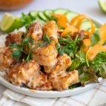 Bang bang chicken served over rice with mixed greens, cucumber slices, carrot ribbons, cilantro, and lime wedges on a white plate.