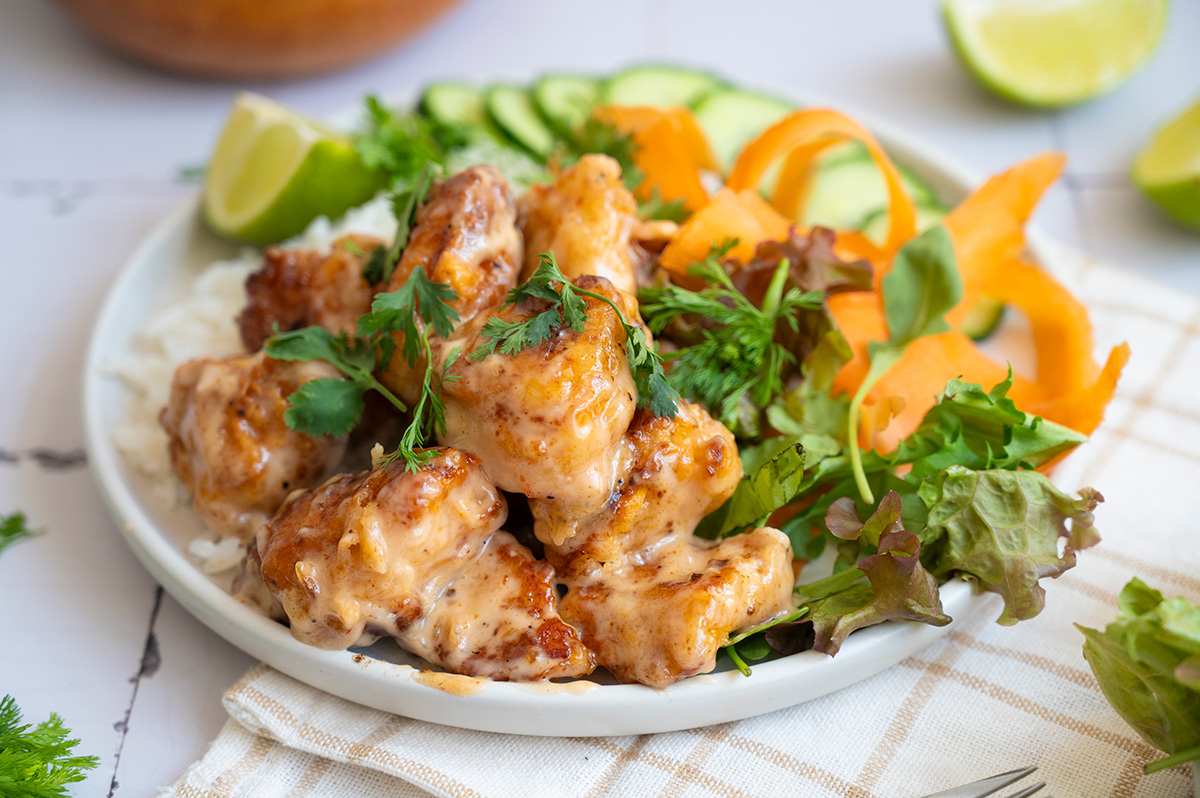 Bang bang chicken served over rice with mixed greens, cucumber slices, carrot ribbons, cilantro, and lime wedges on a white plate.