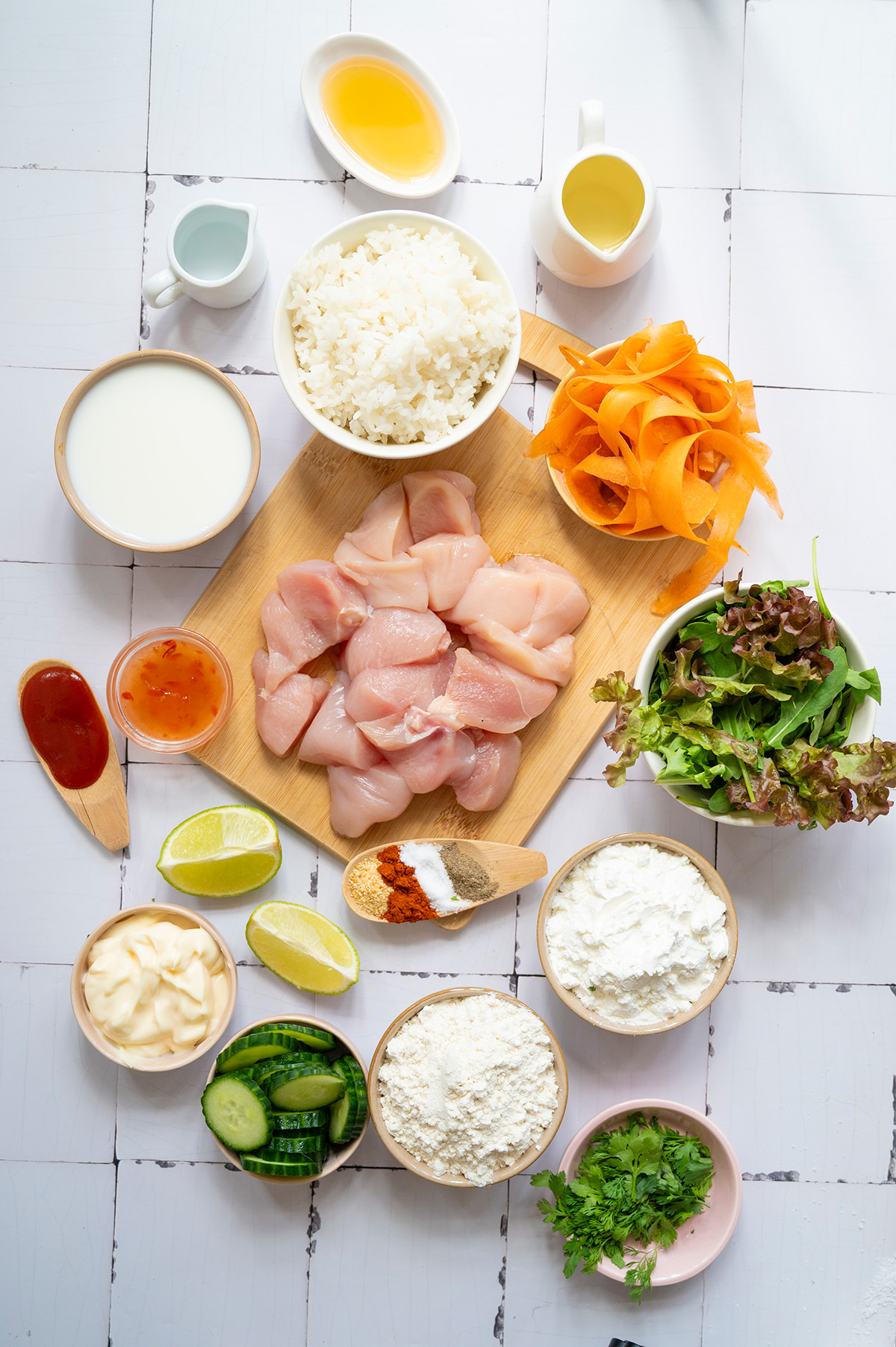 Overhead view of all ingredients for bang bang chicken and rice bowls, including chicken pieces, rice, buttermilk, sweet chili sauce, sriracha, flour, cornstarch, cucumbers, carrot ribbons, greens, spices, lime, and cilantro.