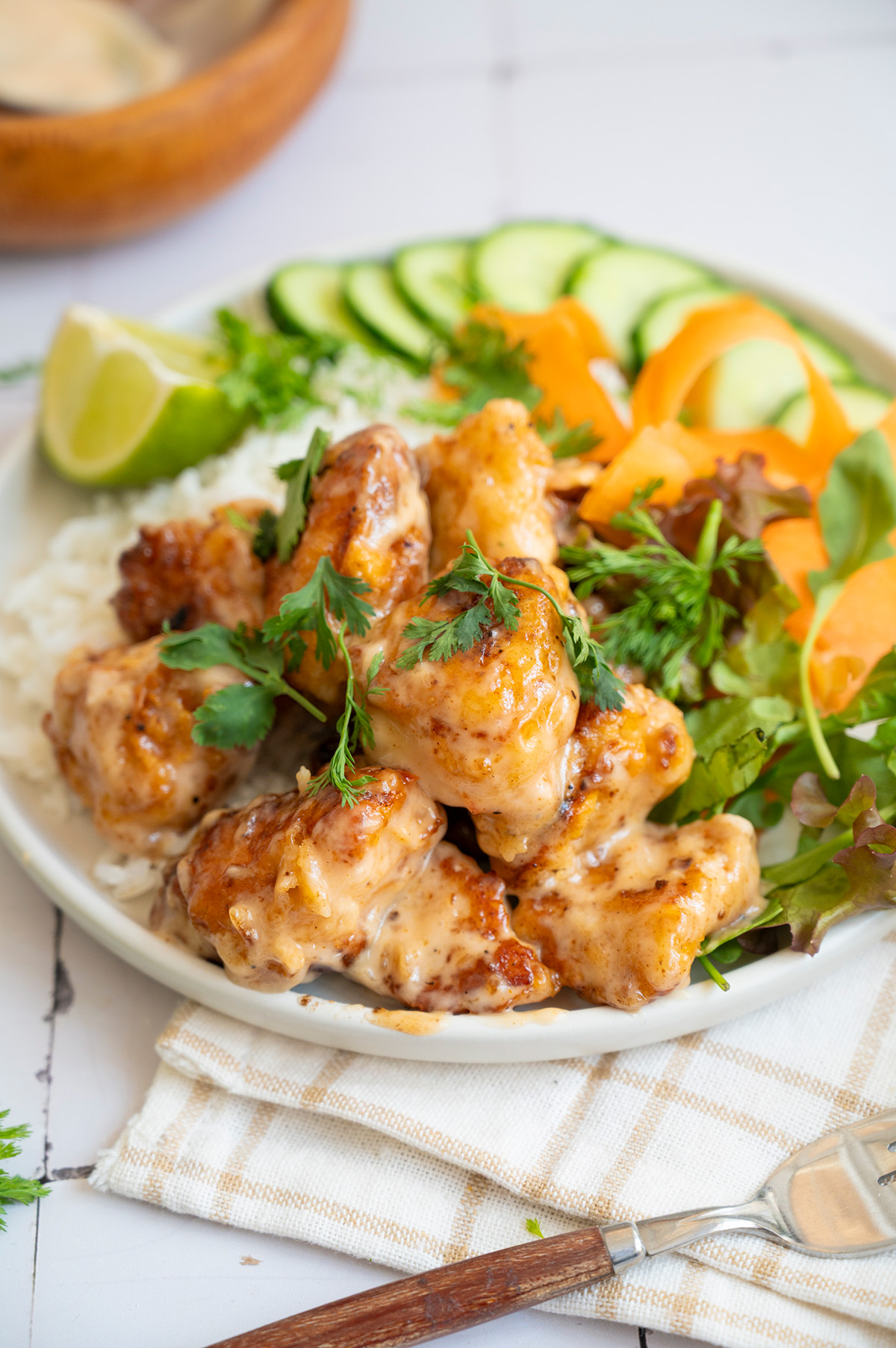 Close-up of bang bang chicken rice bowl with crispy chicken coated in sauce, fresh greens, rice, cucumber, carrot ribbons, and lime.
