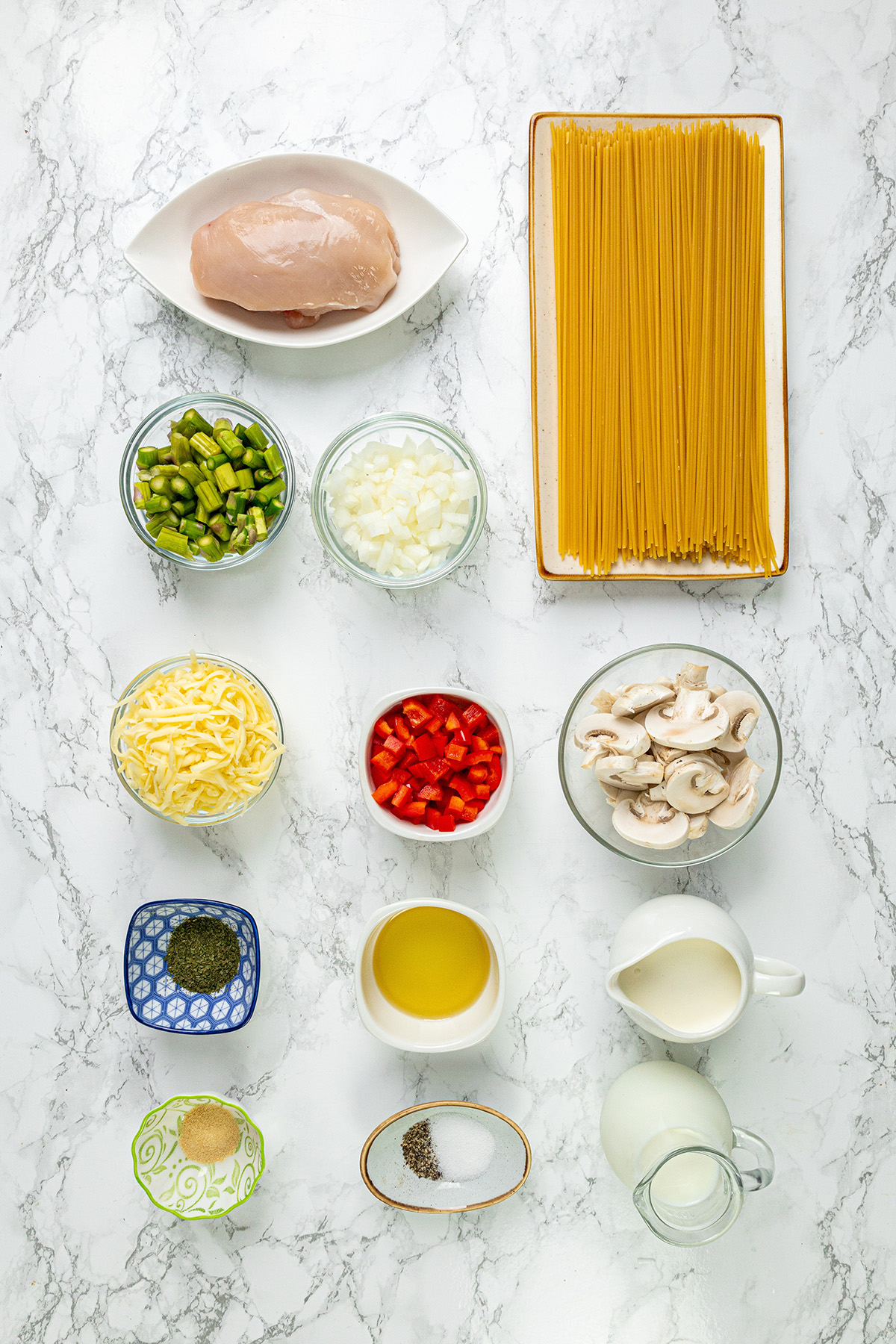 Flat-lay of ingredients for chicken mushroom alfredo pasta including spaghetti, chicken breast, mushrooms, asparagus, bell pepper, onion, mozzarella, olive oil, milk, cream, parsley, and spices.