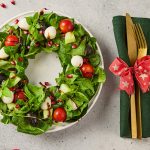 Festive Christmas wreath salad on a plate surrounded by holiday décor, served with a green napkin and gold flatware tied with a red ribbon.