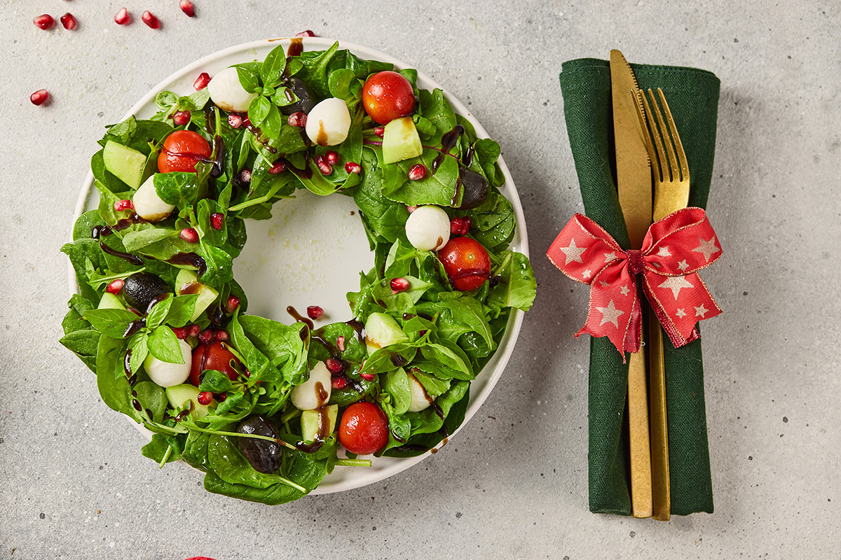Festive Christmas wreath salad on a plate surrounded by holiday décor, served with a green napkin and gold flatware tied with a red ribbon.