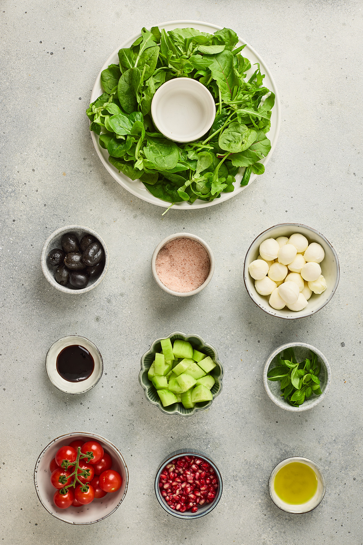 Flat lay of ingredients for a Christmas wreath salad including mixed greens, cherry tomatoes, mozzarella balls, cucumbers, black olives, basil, pomegranate seeds, olive oil, and balsamic glaze.