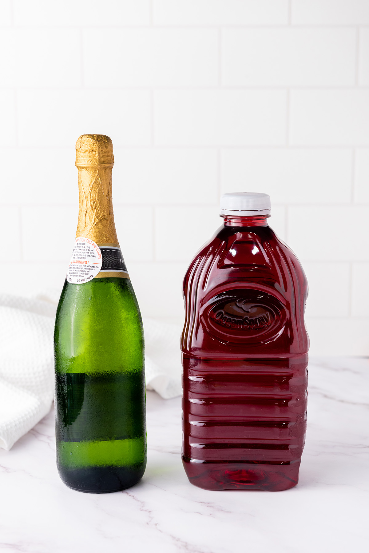 Chilled bottle of brut champagne and a bottle of cranberry juice on a white marble surface.
