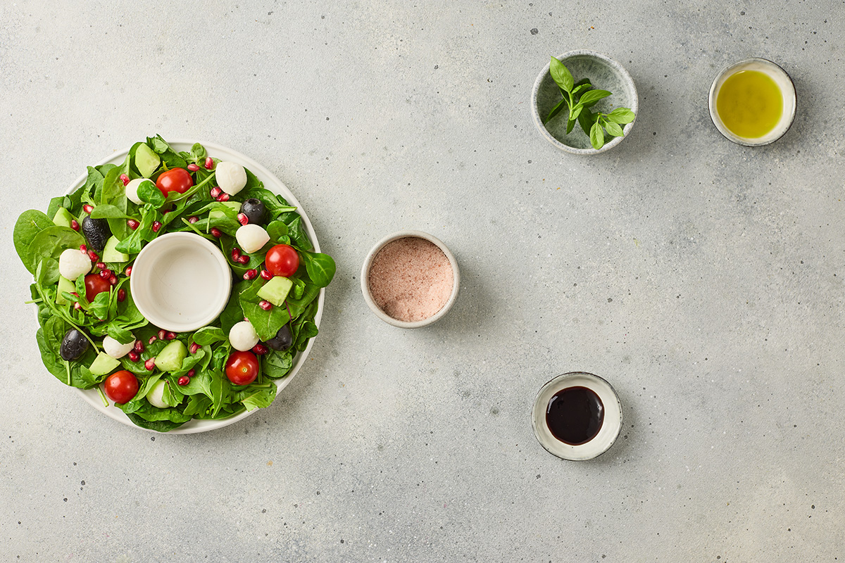 Partially assembled Christmas wreath salad with greens topped with tomatoes, mozzarella balls, olives, pomegranate seeds and cucumbers, with ingredients placed around the plate.